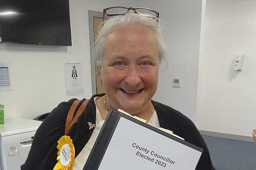 A white woman with glasses on her head, wearing a black cardigan, white top, and a yellow rosette. She is holding a folder saying 'County Councillor Elected 2023 - Welcome Pack - Woodbridge Division'