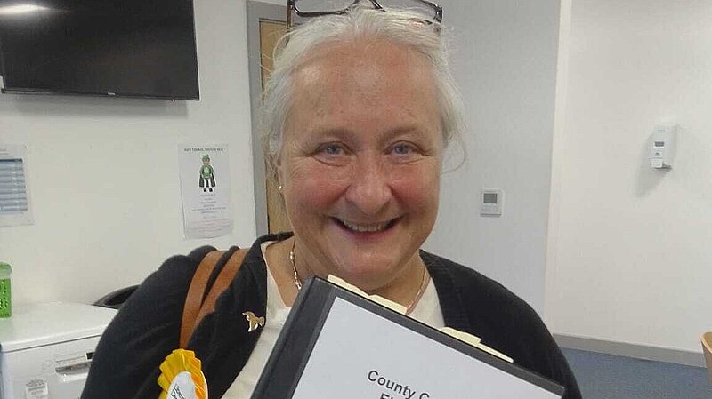 A white woman with glasses on her head, wearing a black cardigan, white top, and a yellow rosette. She is holding a folder saying 'County Councillor Elected 2023 - Welcome Pack - Woodbridge Division'